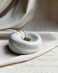 A pair of gold earrings sits in the smooth, round Sumo Pocket Concrete Bowl by Faire, placed on a textured stone surface with a beige fabric draped behind.