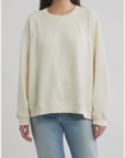 A person wearing The Rehearsal Sweatshirt in a loose, cream-colored raglan-sleeve style by The Great Inc., paired with light blue jeans, stands against a plain, light background. The person's face is partially out of frame.
