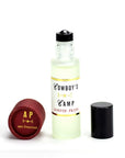 A small frosted glass bottle of Cowboy's Camp Perfume Oil by Faire stands upright, its black cap aside. A red cap with "AP san francisco" in gold lettering sits nearby, completing this unique fragrance’s presentation.