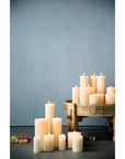 Creative Co-op Unscented Pillar Candles in varying heights, all lit, are arranged on the floor and a wooden pedestal with dried flowers nearby against a pale blue backdrop. Burn time: up to 145 hours.