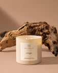 A hand poured 14oz. white luxury candle labeled "Rouge" from Faire rests on a beige surface with textured driftwood, evoking the sophisticated aroma of Baccarat Rouge 540 in a warm, neutral setting.