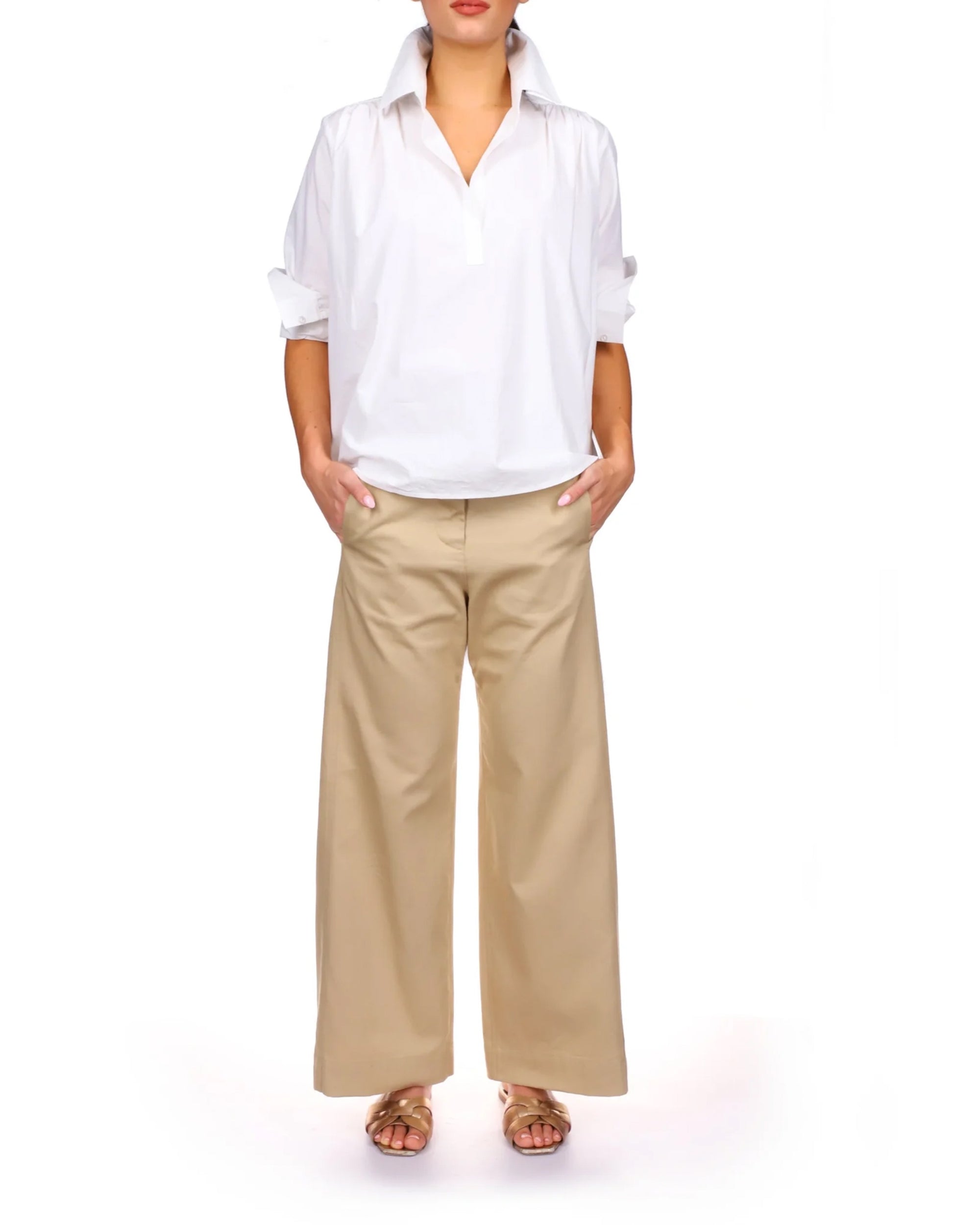 A person stands with hands in pockets, wearing the Celeste Parachute by A Shirt Thing—a 100% cotton white button-up shirt with rolled sleeves—paired with beige wide-leg pants and brown sandals against a plain white background.