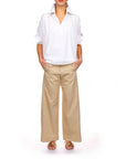 A person stands with hands in pockets, wearing the Celeste Parachute by A Shirt Thing—a 100% cotton white button-up shirt with rolled sleeves—paired with beige wide-leg pants and brown sandals against a plain white background.