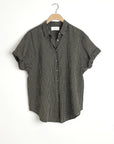 The Xirena Channing Shirt S25, a short-sleeved black and white checkered cotton chambray piece, hangs on a wooden hanger against a plain white wall.