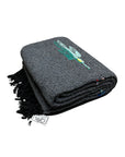 A folded Charcoal Thunderbird Baja Blanket by Faire features a dark gray woven design with black fringe, subtle colorful patterns on top, and a visible branded tag highlighting its handmade quality in Mexico.