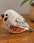 Gray hand-painted carved wood bird by Faire on a brown surface with nature-inspired background accents.