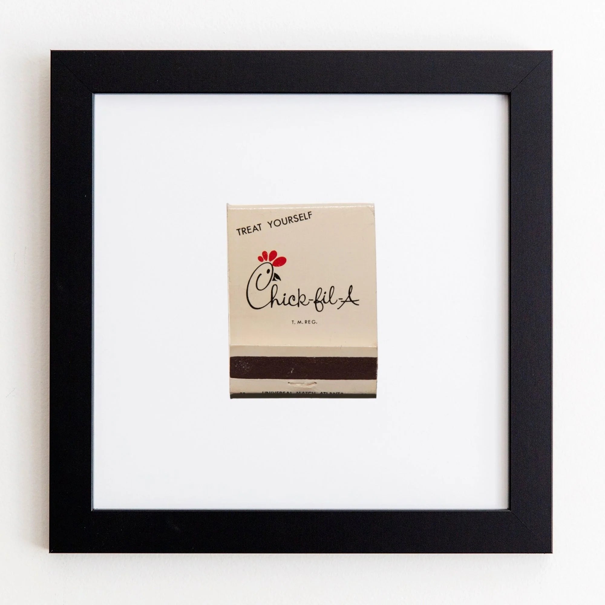 A framed Match South "Treat Yourself" gift card mounted on a white wall. The card features the Chick-fil-A logo and a small red heart above the acrylic text.