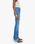 A woman stands in profile against a white background wearing a white sleeveless top, blue high-waisted bootcut jeans made from recycled cotton with frayed hems, a gold watch, and The Chomp Heel Chew by Mother—black pointed-toe heels with an ankle strap.