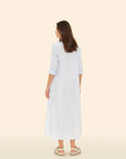 A person with long hair faces away, wearing the Boden Dress by Xirena in light blue cotton poplin with rolled-up sleeves. They pair it effortlessly with beige sandals against a solid, light beige background.