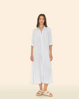 A person in a light blue Boden Dress from the brand Xirena stands against a beige background. They wear beige sandals and a multicolored necklace, with hands by their sides. The classic, tailored fit cotton poplin shirtdress falls below the knees.