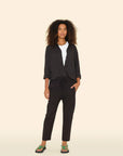 A woman with curly hair wears the Xirena Draper Pant in black, paired with a black lightweight cotton chambray button-up over a white top and black sandals with green straps, standing against a plain beige background.