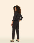 A woman with curly hair stands sideways and looks over her shoulder, wearing a black long-sleeve shirt, Xirena's Draper Pant in black with side seam pockets, and green sandals—all set against a plain beige background.