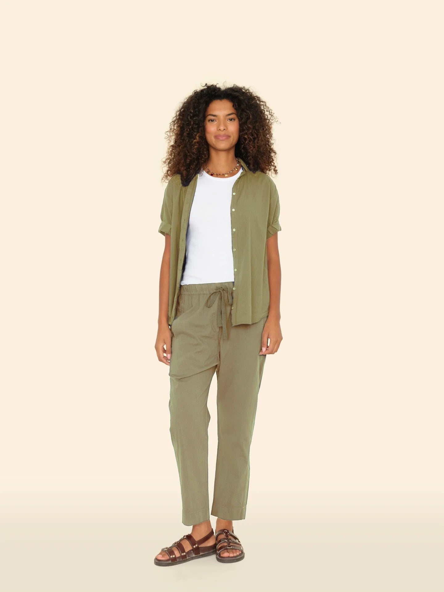 A person with curly hair stands against a plain background, wearing an olive green open shirt over a white top and Xirena's Draper Pant in olive green, crafted from lightweight cotton chambray. They complete the look with brown sandals and gaze at the camera neutrally.