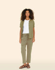 A person with curly hair stands against a plain background, wearing an olive green open shirt over a white top and Xirena's Draper Pant in olive green, crafted from lightweight cotton chambray. They complete the look with brown sandals and gaze at the camera neutrally.
