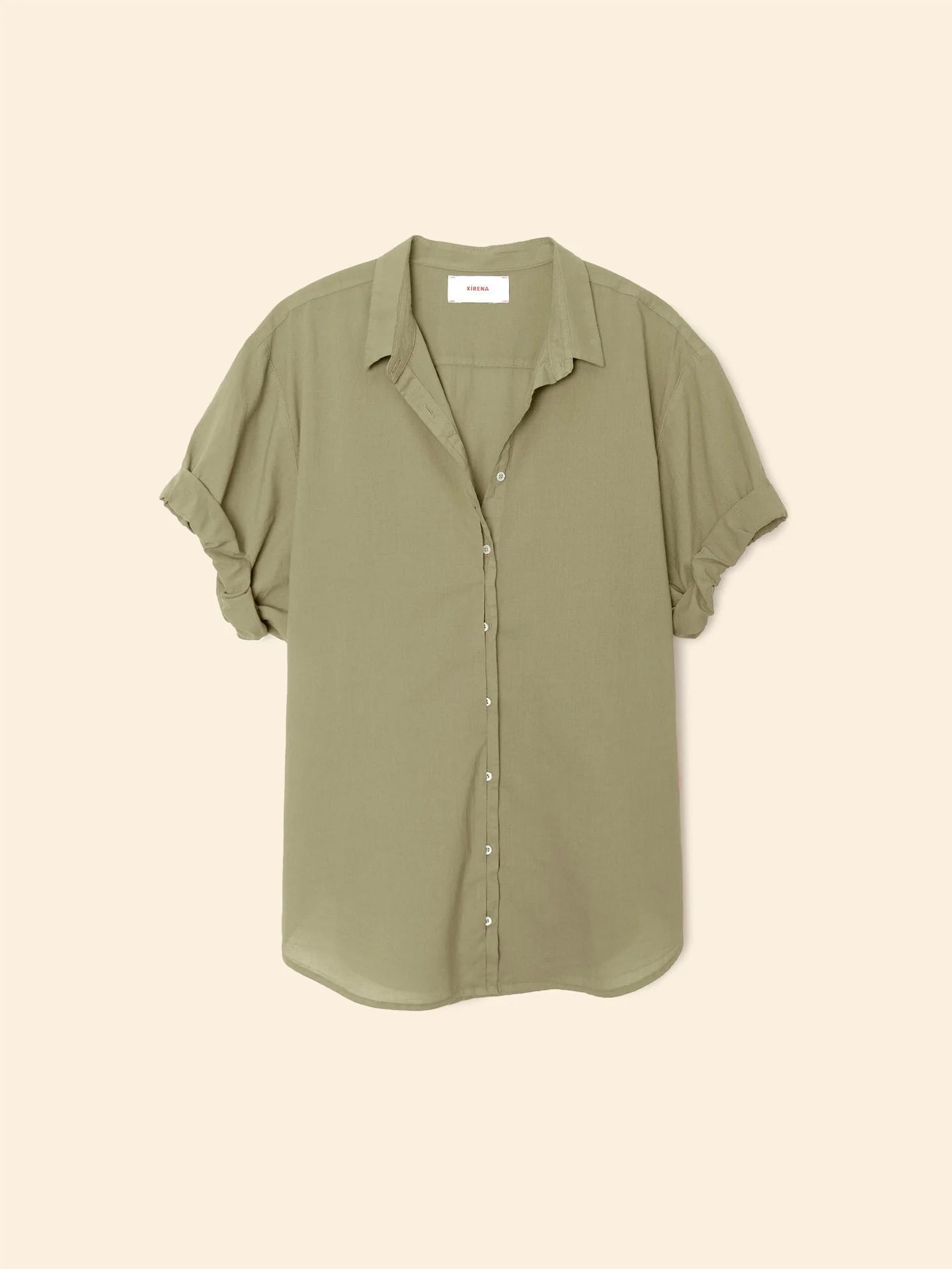 The Xirena Channing Shirt S25 is a light olive green, short-sleeve cotton chambray shirt with rolled cuffs, relaxed fit, collar, and a small white neckline label, shown laid flat on a beige background.