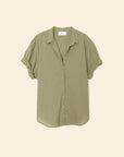 The Xirena Channing Shirt S25 in light olive green cotton chambray features rolled short sleeves, displayed against a beige background—ideal for a casual "boy meets girl" vibe.