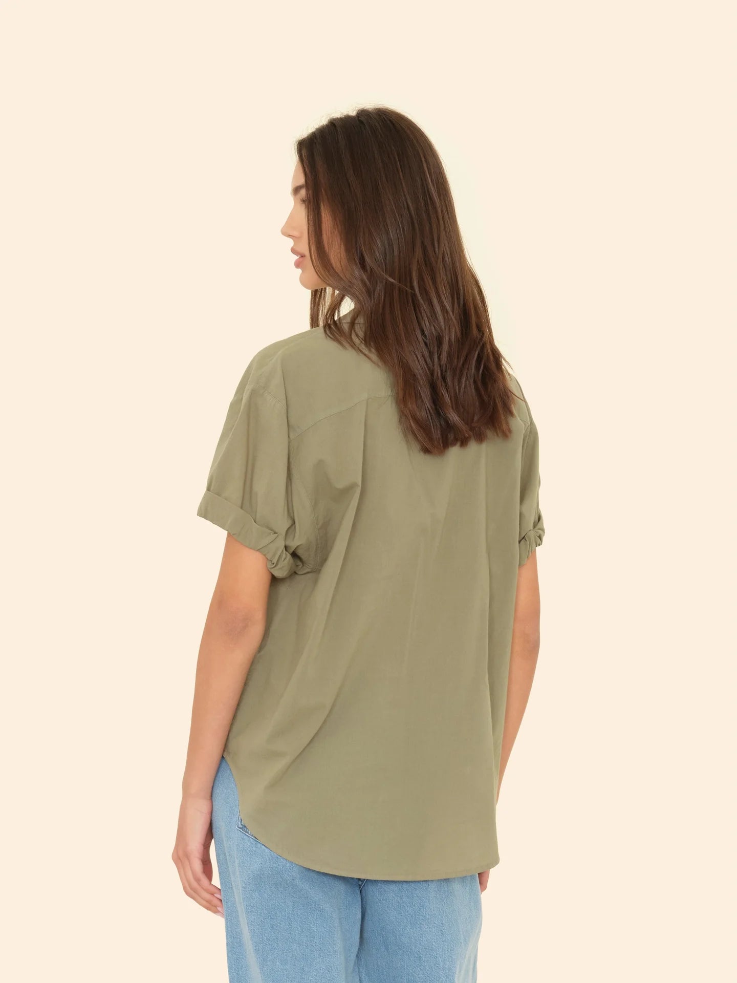A woman with long brown hair, wearing a short-sleeved olive green Channing Shirt S25 by Xirena and light blue wash jeans, stands facing away from the camera against a plain beige background.