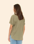 A woman with long brown hair wears the Xirena Channing Shirt S25 in olive green cotton chambray, paired with light blue jeans. She stands with her back to the camera against a beige background, embodying a "boy meets girl" vibe.