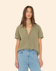 A woman wears the Xirena Channing Shirt S25—an olive green, short-sleeve cotton chambray button-up—paired with high-waisted light blue jeans and a beaded necklace, creating a "boy meets girl" vibe against a beige background.