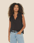 A person with curly hair is wearing the Bex Top by Xirena, a sleeveless black top with ruffled shoulders and a V-neck, paired with light blue jeans. They have a necklace and are standing against a plain, light-colored background while looking forward.