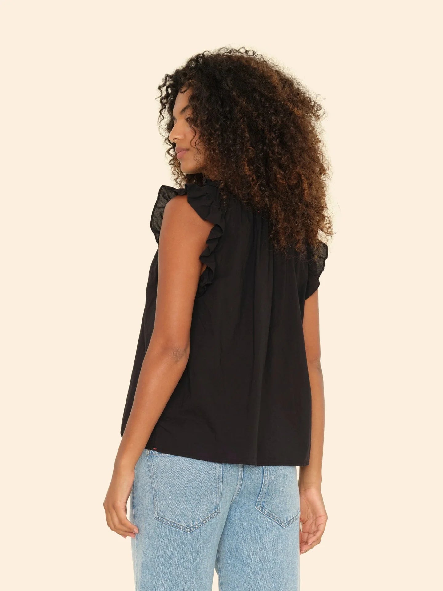A person with curly hair wearing the Xirena Bex Top, a sleeveless black piece with ruffle detailing and a V-neck, paired with light blue jeans, stands looking to the side against a plain beige background.