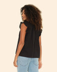 A person with curly hair wearing the Xirena Bex Top, a sleeveless black piece with ruffle detailing and a V-neck, paired with light blue jeans, stands looking to the side against a plain beige background.