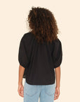 A person with curly hair is seen from the back wearing the Xirena Jules Top, a black cotton poplin blouse with bubble sleeves, and light blue jeans against a beige background.