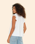 A person with curly hair stands sideways in the Bex Top by Xirena, featuring sleeveless white cotton poplin with ruffled shoulders, paired with light blue jeans against a soft beige background.