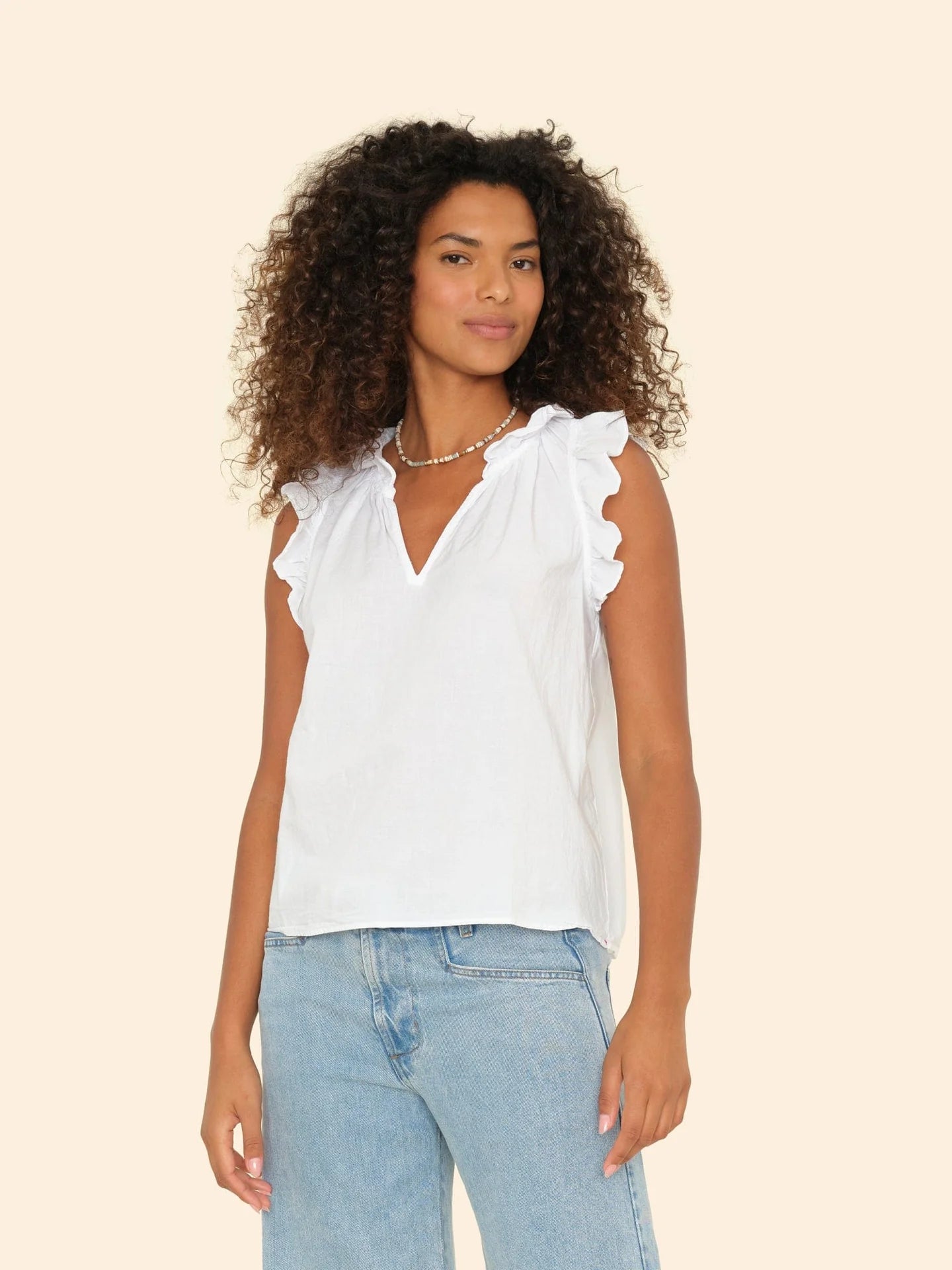 A person with curly hair wears the Bex Top, a white sleeveless cotton poplin piece from Xirena, featuring ruffle details on the V-neck. They're paired with light blue jeans and posed against a plain light beige background, exuding a relaxed confidence.