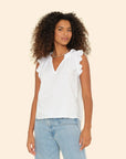 A person with curly hair wears the Bex Top, a white sleeveless cotton poplin piece from Xirena, featuring ruffle details on the V-neck. They're paired with light blue jeans and posed against a plain light beige background, exuding a relaxed confidence.