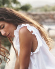 A woman with long, wavy brown hair is outdoors in soft, natural light, wearing the Bex Top by Xirena. It's a sleeveless white blouse made of cotton poplin with ruffle detailing and a V-neck, surrounded by blurred greenery and a stone structure.