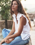 A woman with long brown hair sits casually outdoors in a sleeveless Bex Top by Xirena, featuring ruffle detailing, paired with blue jeans and a blue necklace. Her expression is relaxed amidst a natural backdrop of trees and soft lighting.