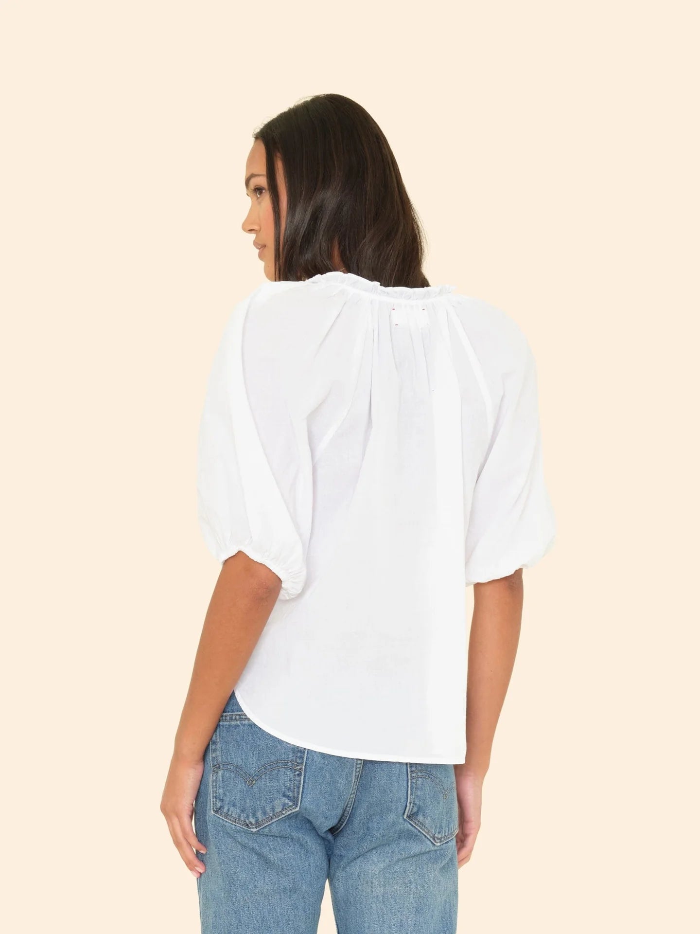 A woman with medium-length dark hair is shown from the back, modeling the Xirena Jules Top—an airy white cotton poplin blouse with bubble sleeves—paired with light blue jeans against a plain beige background.