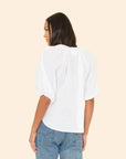 A woman with medium-length dark hair is shown from the back, modeling the Xirena Jules Top—an airy white cotton poplin blouse with bubble sleeves—paired with light blue jeans against a plain beige background.