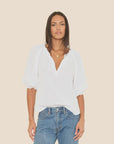 A woman with straight, shoulder-length hair and a gold necklace wears the Xirena Jules Top—a loose-fitting white cotton poplin blouse with bubble sleeves—paired with blue jeans, standing against a plain beige background.