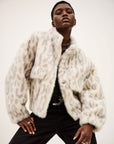 A person poses confidently in the BA&SH Coat Saros, a loose fit, light-colored cropped faux fur jacket with bold animal print and large buttons, paired with black pants against a plain background.