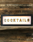 The Faire Lighted Sign is a rectangular, wall-mountable sign displaying "COCKTAILS" in bold black letters, illuminated against a wooden plank wall background.