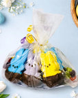 A clear gift bag tied with yellow ribbon holds Faire Easter Dipped Peeps—blue, purple, and yellow bunnies dipped in milk chocolate—surrounded by small candies on a pastel blue surface near flowers and an Easter basket.