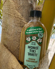 A bottle of Faire Monoi Coco Coconut Oil with a green label and white Tiare flower illustrations rests against a palm tree, set on a sandy beach with lush palm fronds in the background.