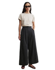 A person stands against a white background with hands in pockets, wearing Rachel Comey’s black Coxsone Pant (organic cotton, wide leg), a beige short-sleeve top, dark sunglasses, a necklace, and sandals.