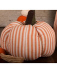 The Faire Pumpkin, featuring soft orange and white vertical stripes with a brown fabric stem, sits on a dark surface for charming farmhouse decor; another plaid pumpkin appears in the background.