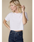 A person with light hair wears The Cropped Perfect Tee by Sold Out NYC—an organic cotton, plain white crop top—with high-waisted dark jeans, standing against a neutral beige background and looking to the side while touching their hair.