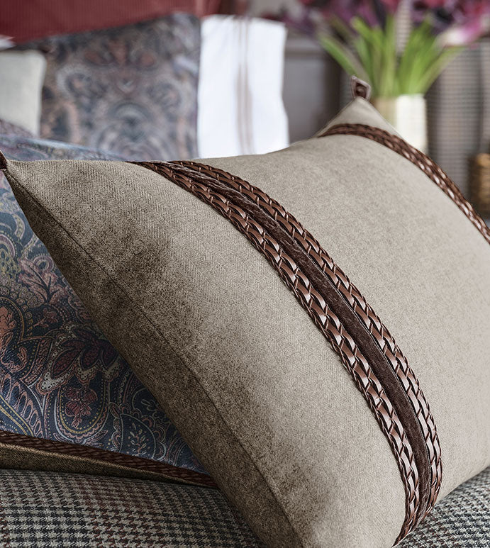 The CRISTOBAL FAUX LEATHER bolster pillow by Eastern Accents, featuring two horizontal woven leather braided accents in beige, rests on a bed with patterned bedding and pillows; a vase of purple flowers sits in the blurred background.
