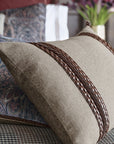 The CRISTOBAL FAUX LEATHER bolster pillow by Eastern Accents, featuring two horizontal woven leather braided accents in beige, rests on a bed with patterned bedding and pillows; a vase of purple flowers sits in the blurred background.