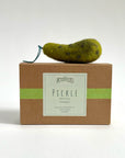A small Felted Ornament by Faire, shaped like a pickle with a green string stem, sits atop a brown box labeled "Pettifelts Pickle" with a green stripe, set against a plain white background.