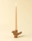 A lit beige taper candle sits in the sculptural Indaba Wren Candle Holder, bird-shaped and set against a light cream background—an ideal rustic accent for your decor.