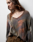 A woman with wavy brown hair looks down while wearing the Riverside Tool & Dye Cashmere V-neck sweater, featuring a blurred multicolor pattern, paired with brown corduroy pants against a plain white background.