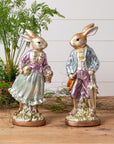 The Faire Vintage Foil Rabbit figurines, dressed in vintage-style clothing, stand together on a wooden surface with greenery—perfect heirloom-inspired decor for charming Easter displays.
