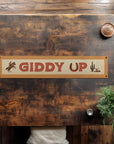 The "Giddy Up" Felt Banner by Faire features a cowboy on horseback and a cactus, adding rustic Wild West charm to your décor. Displayed on a wooden table with vintage accents like a plant and cup, two hands rest on opposite sides.