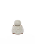 The Alabaster White Stash Jar by Faire, a small round stoneware jar with a matching lid in light beige and dark brown flecks, finished with a matte glaze, is centered on a white background.